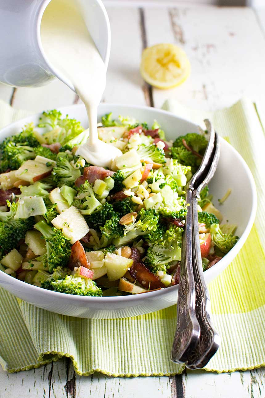 Someone pouring dressing over a bowl of broccoli, apple and bacon salad on a green cloth and with serving spoons in it.