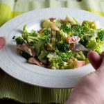 Someone eating from a white bowl of healthy broccoli, apple and bacon salad.