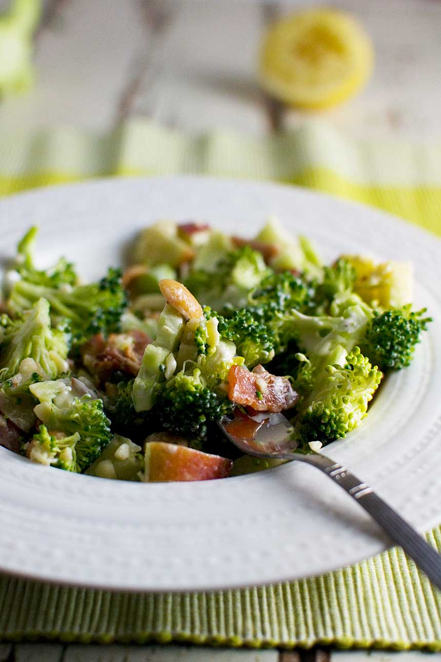 Closeup of a white bowl of broccoli, apple and bacon salad with a spoon in it.
