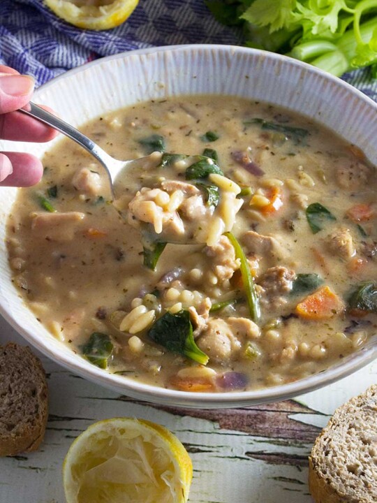 Closeup of someone eating lemon chicken orzo soup out of a white bowl with a spoon with lemons and other ingredients in the background