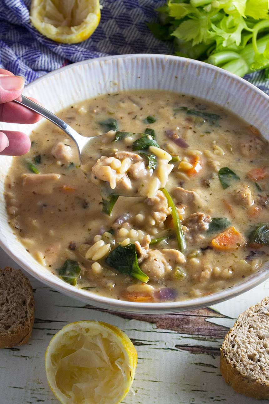 Closeup of someone eating lemon chicken orzo soup out of a white bowl with a spoon with lemons and other ingredients in the background