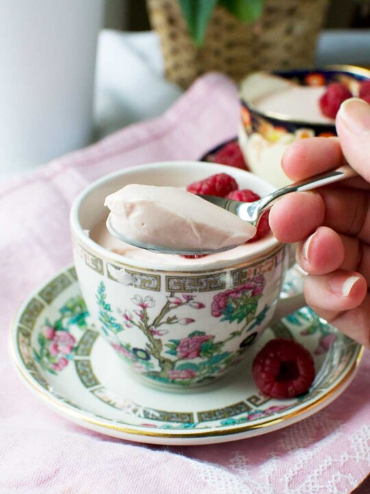 Someone spooning raspberry Greek yogurt panna cotta out of a pretty tea cup on a pink cloth.