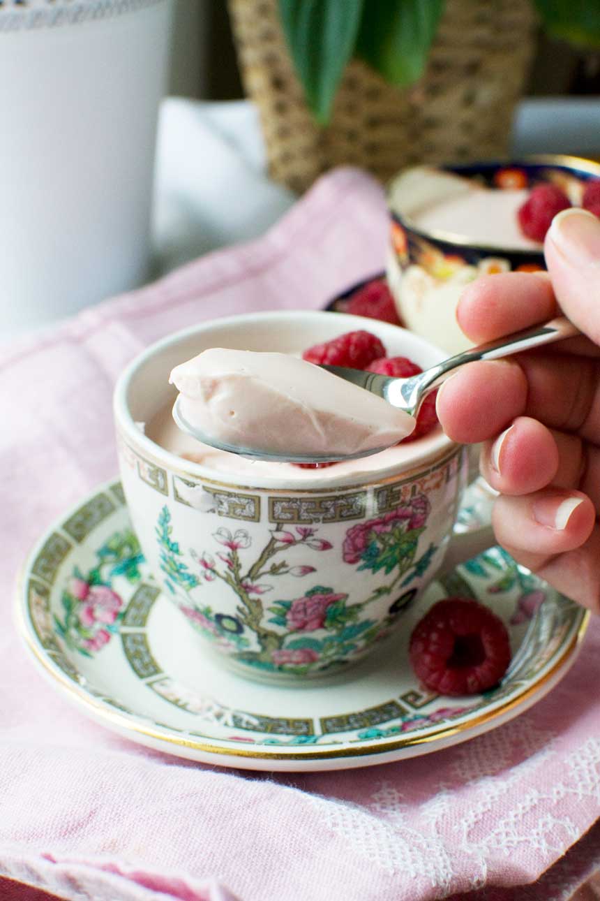 Someone spooning raspberry Greek yogurt panna cotta out of a pretty tea cup on a pink cloth.