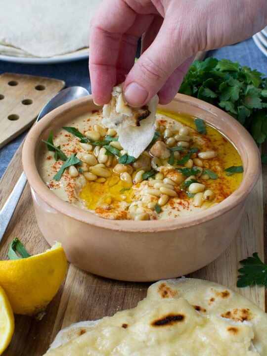 Someone dipping bread into hummus in a terracotta pot with bread and lemon around it.