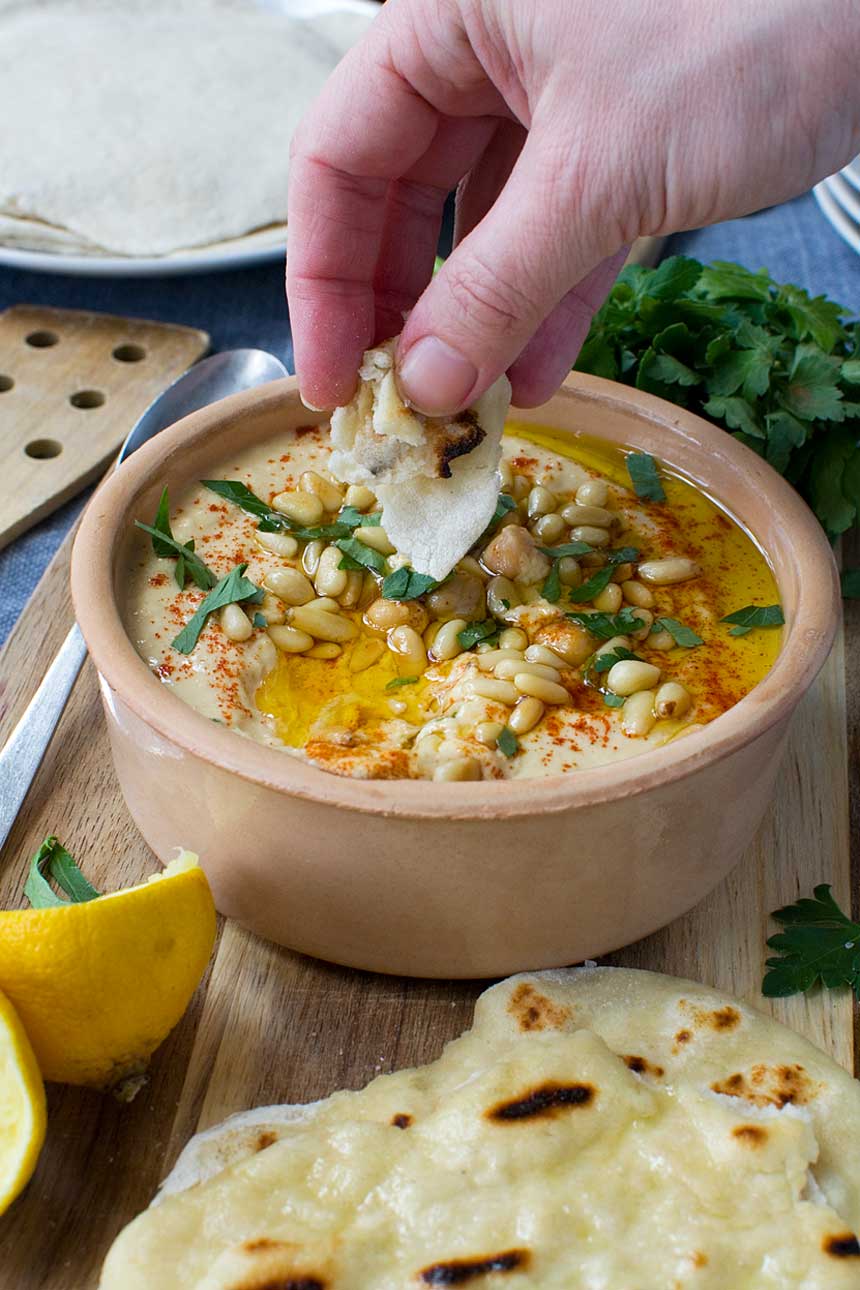 Someone dipping bread into hummus in a terracotta pot with bread and lemon around it.