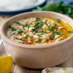 A brown ceramic pot with hummus in it sprinkled with pine nuts, olive oil and herbs and with ingredients scattered around.