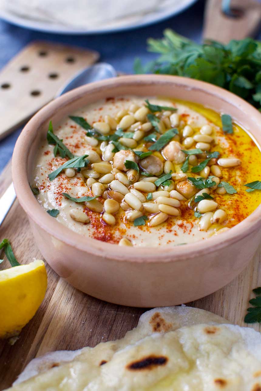 Quick hummus in a brown ceramic pot with pine nuts, paprika, olive oil and herbs on top.