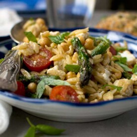 A closeup of a blue bowl of asparagus, tomato and chickpea orzo salad with a spoon across the top.