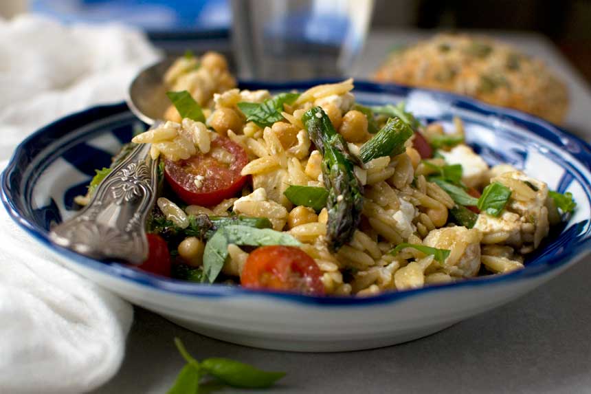 A closeup of a blue bowl of asparagus, tomato and chickpea orzo salad with a spoon across the top.