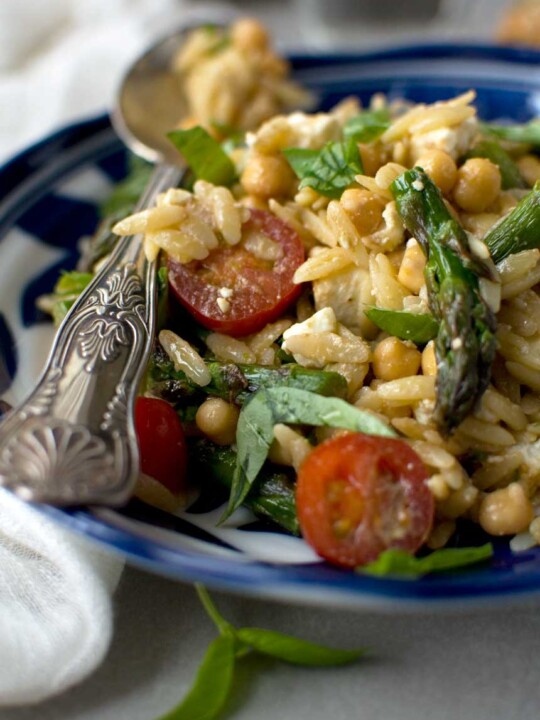 Closeup of an Asparagus, tomato and feta orzo salad in a blue patterned dish and with a patterned serving spoon.