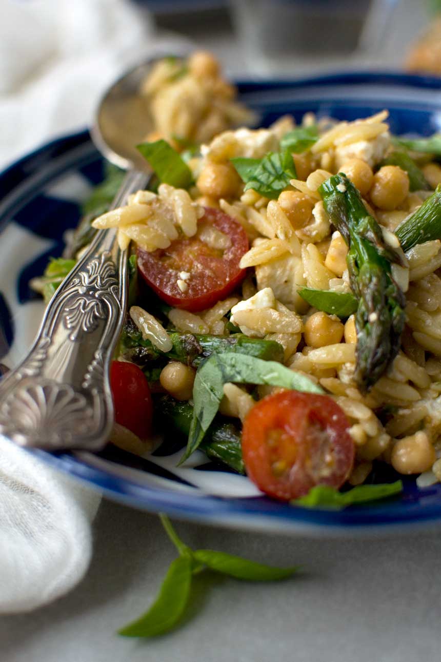 Closeup of an Asparagus, tomato and feta orzo salad in a blue patterned dish and with a patterned serving spoon.