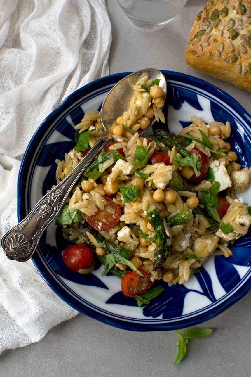 Asparagus, tomato and feta orzo salad in a blue patterned bowl and with a patterned spoon in it.