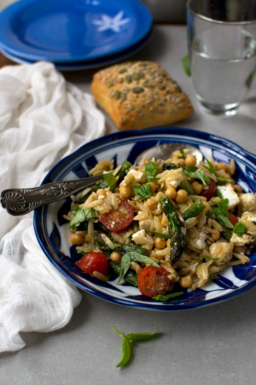 Bowl of asparagus, tomato and feta orzo salad with a spoon in it and with bread in the background.