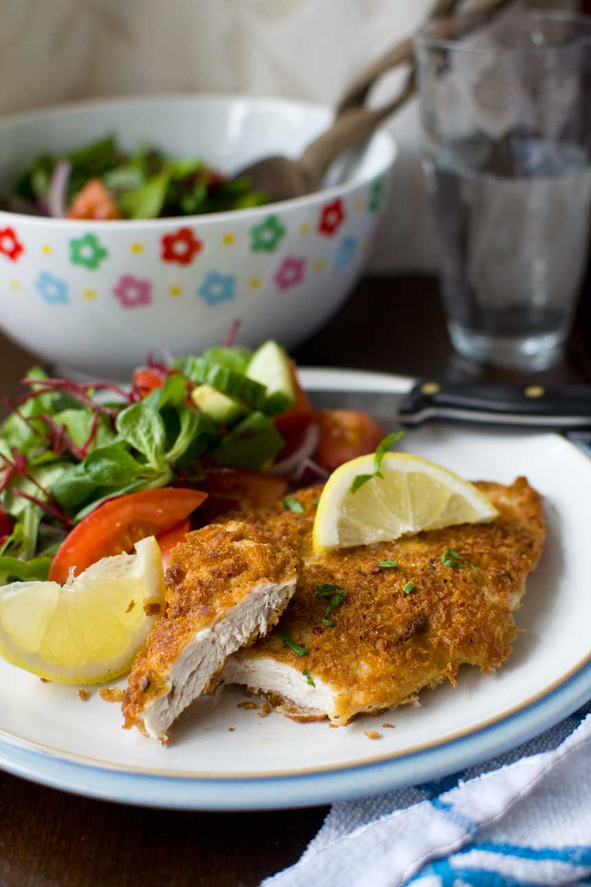 A piece of crispy mustard parmesan chicken on a white plate with lemon segments and salad and with a pretty bowl in the background.