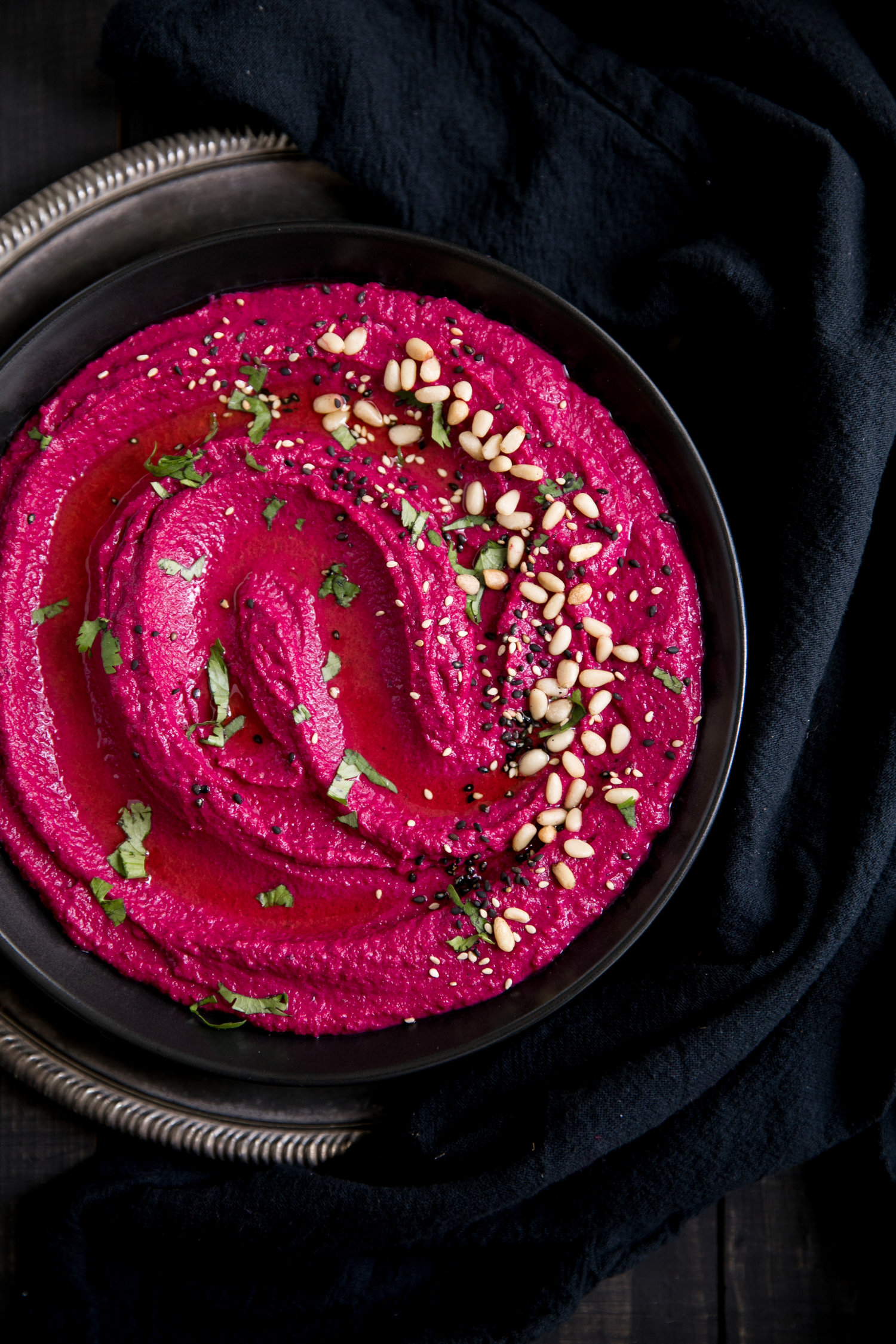 A close up of the top of a bowl of purple hummus with nuts on top.