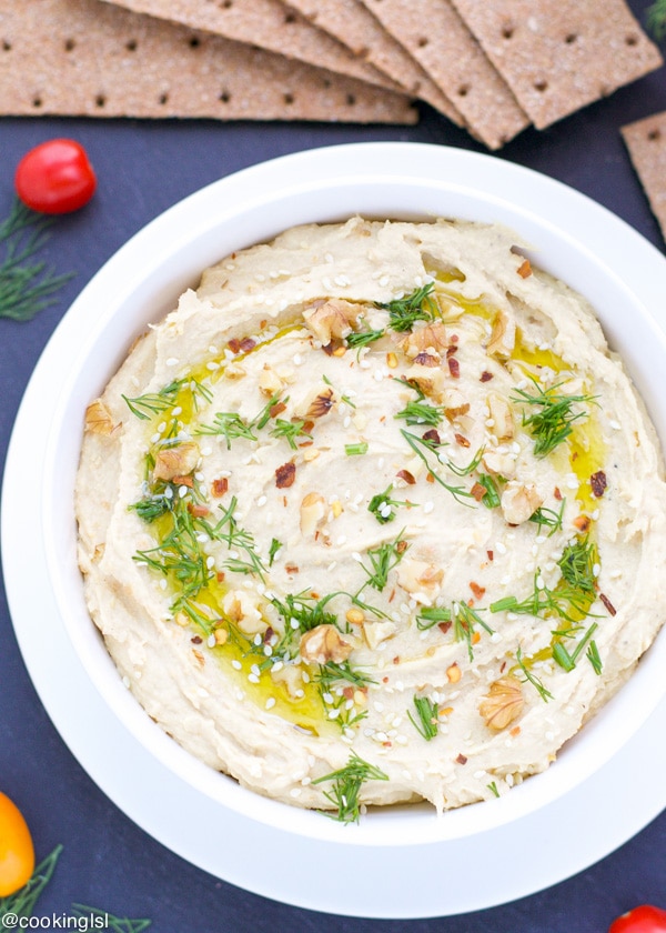 A bowl of Creamy roasted garlic and lemon hummus with fresh herbs scattered over it from above.