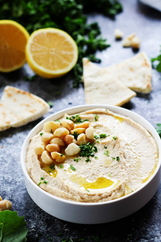 A bowl of hummus with chickpeas and lemon on top of it with tortilla bread and lemons in the background.