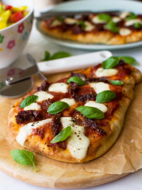 A sun dried tomato and bocconcini naan pizza on a cutting board with dishes in the background