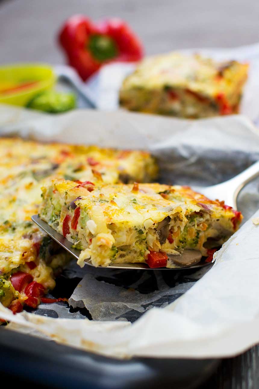 Someone lifting a piece of sheet pan frittata out of a baking dish with a slice with more in the background.