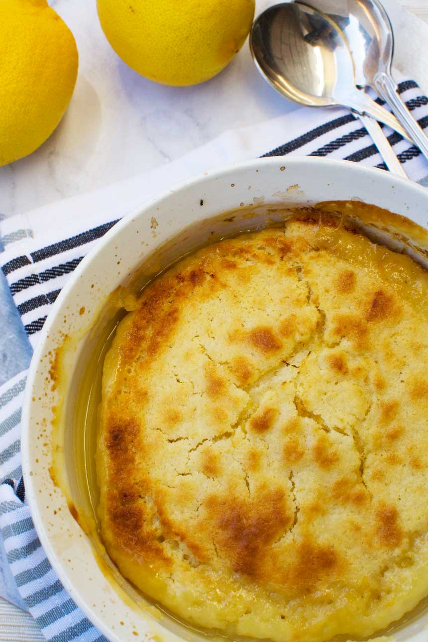 A souffle dish of easy \'magic\' lemon pudding from above on a marble background with lemons and a blue striped tea towel under the dish.