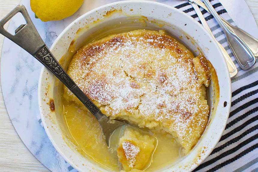 A souffle dish of easy \'magic\' lemon pudding from above with a spoon in it on a marble background with lemons and a blue striped tea towel.