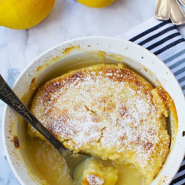 A souffle dish of easy 'magic' lemon pudding from above with a spoon in it on a marble background with lemons and a blue striped tea towel.