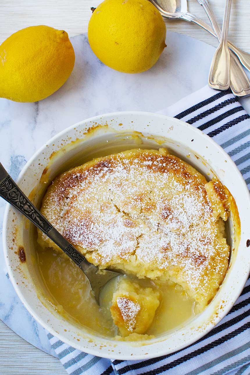 A souffle dish of easy \'magic\' lemon pudding from above with a spoon in it on a marble background with lemons and a blue striped tea towel.