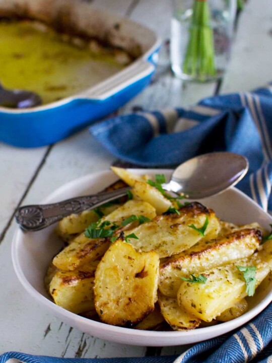 A white dish of cooked Greek potatoes with fresh herbs on top with a spoon in it and with a blue tea towel and baking dish in the background.
