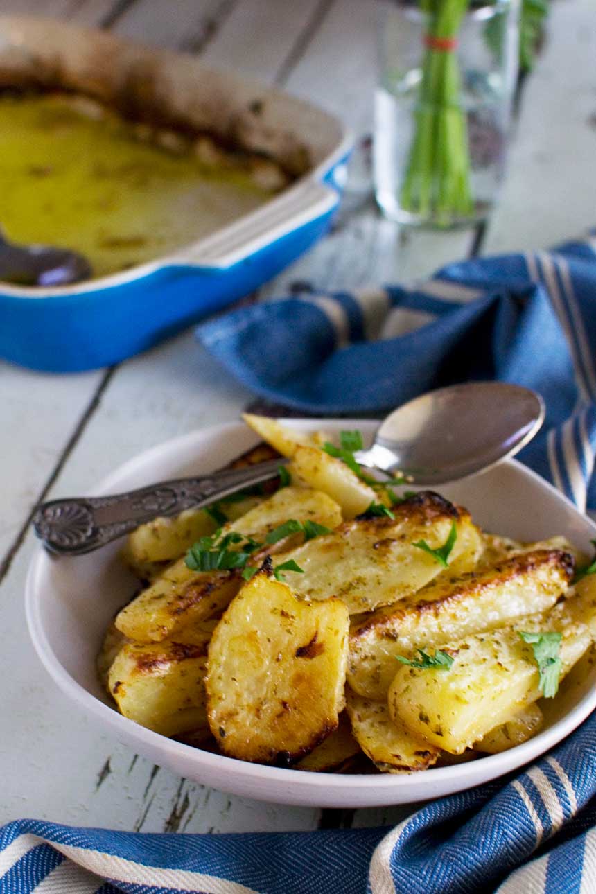 A white dish of cooked Greek potatoes with fresh herbs on top with a spoon in it and with a blue tea towel and baking dish in the background.