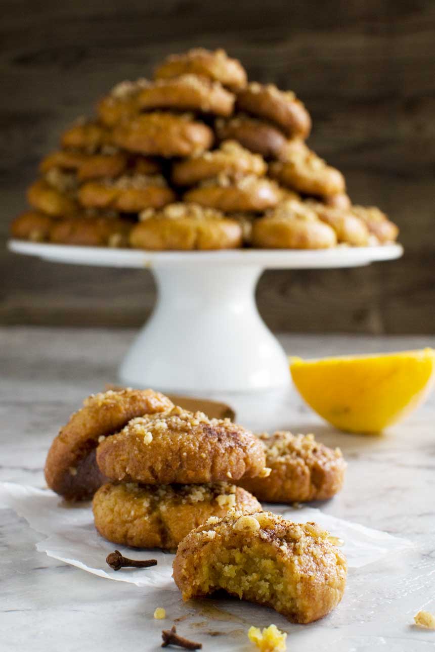 A Greek Christmas honey cookie with a bite out of it with more cookies behind it and a pyramid of them on a cake stand behind.
