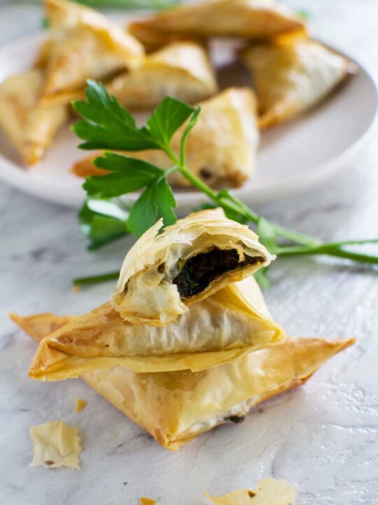 A small stack of crispy mushroom filo triangles on a marble background with more on a plate in the background.