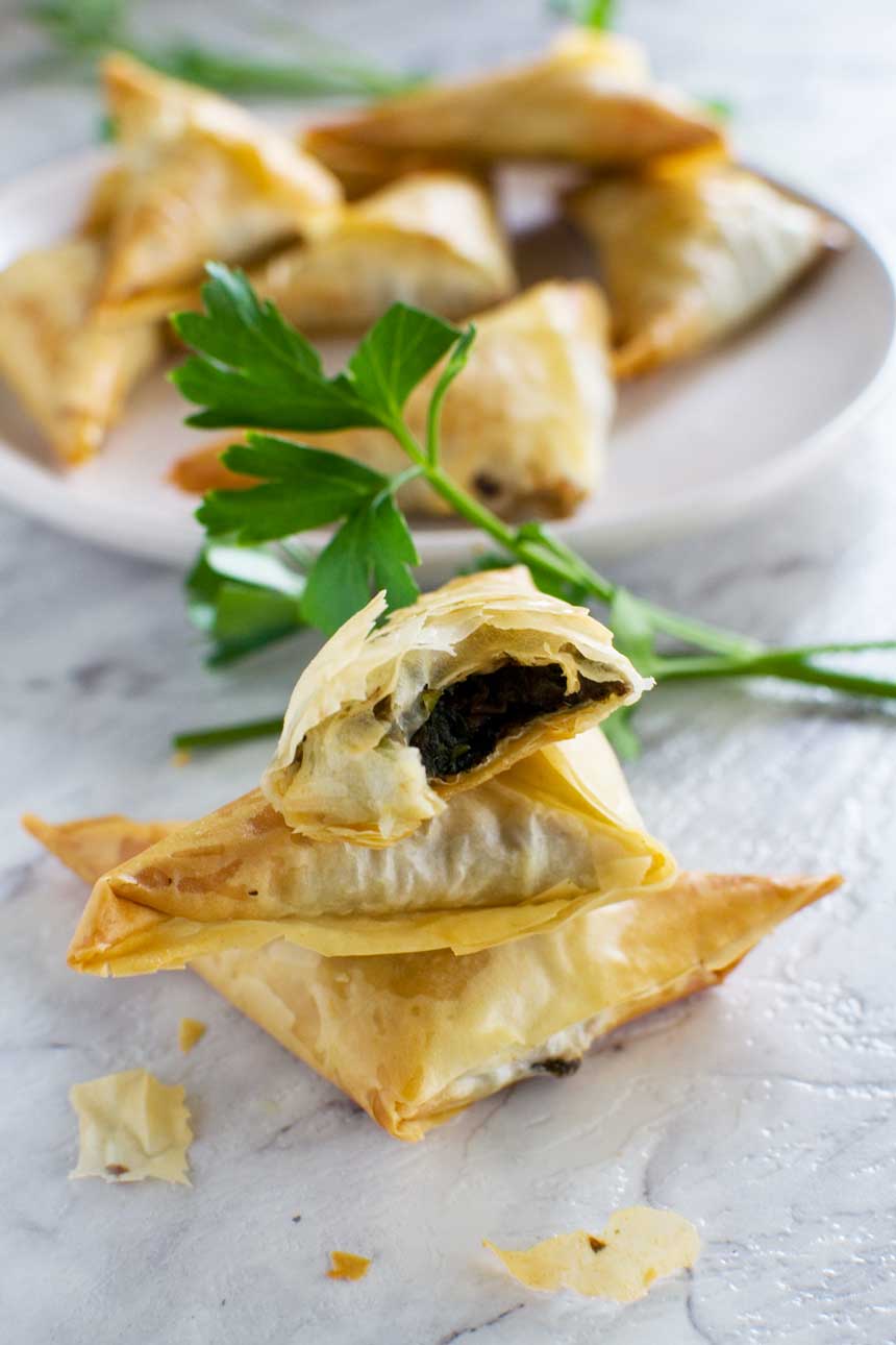 A small stack of crispy mushroom filo triangles on a marble background with more on a plate in the background.