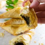 Someone holding a crispy mushroom filo triangle with a bite out of it, a stack of them in the background