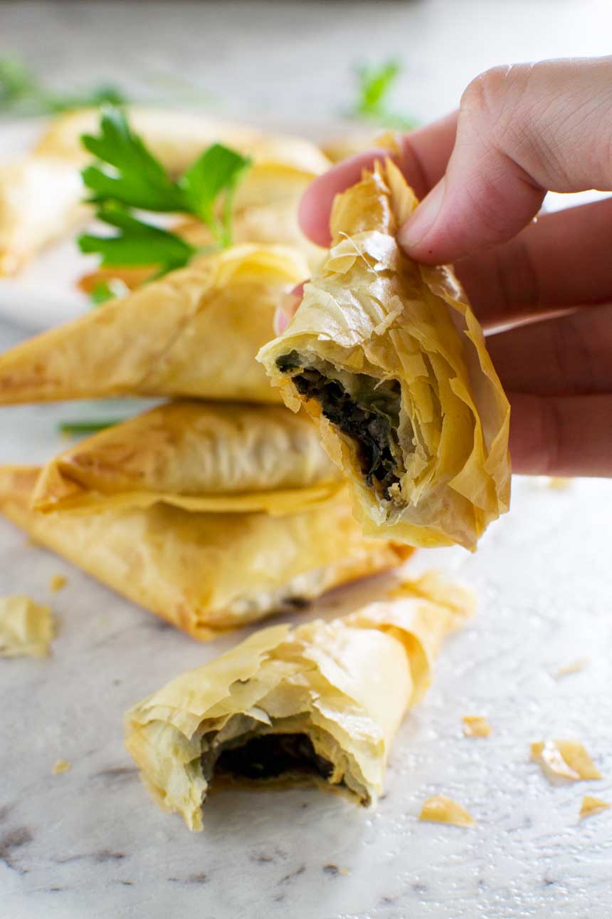 Someone holding a crispy mushroom filo triangle with a bite out of it, a stack of them in the background