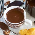 A cup of thick hot chocolate in a brown cup with a spoon and ingredients and kitchen equipment in the foreground and background.