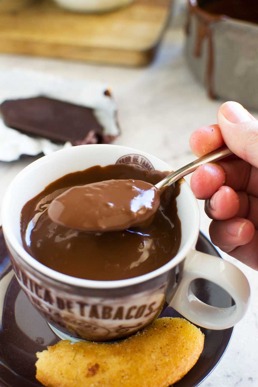 Someone lifting up a spoon of thick hot chocolate from a brown coffee cup of chocolate on baking paper.