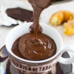 Someone pouring thick hot chocolate into a brown coffee cup with ingredients in the background.