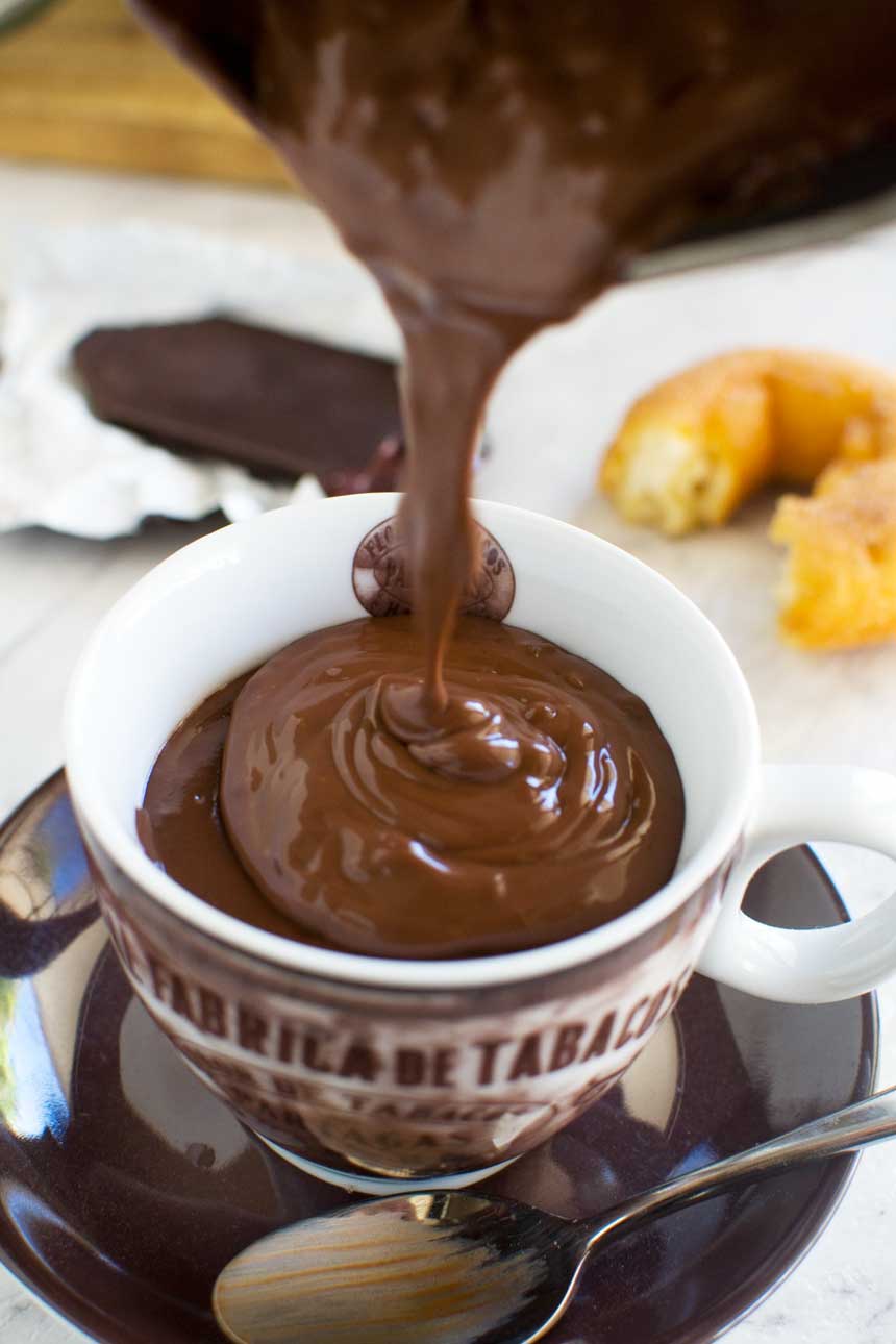 Someone pouring thick hot chocolate into a brown coffee cup with ingredients in the background.