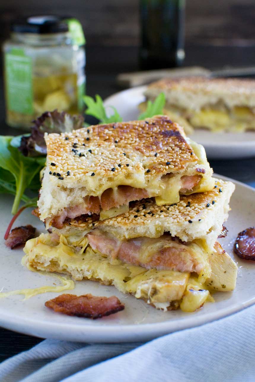 Artichoke and bacon grilled cheese sandwich on a white plate with another sandwich and ingredients in the background