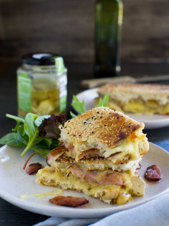 An Artichoke and bacon grilled cheese sandwich stacked up on a white plate with ingredients in the background.