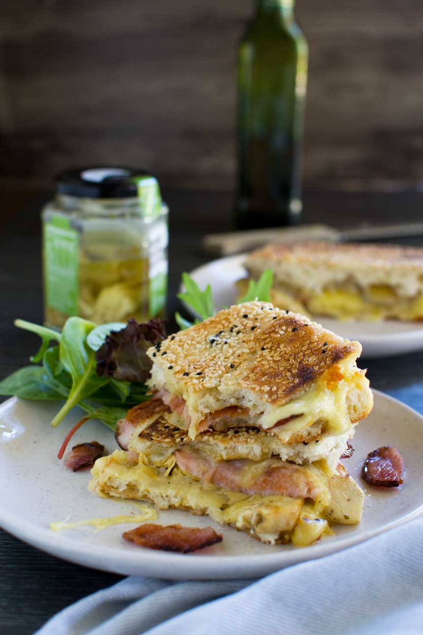 An Artichoke and bacon grilled cheese sandwich stacked up on a white plate with ingredients in the background.