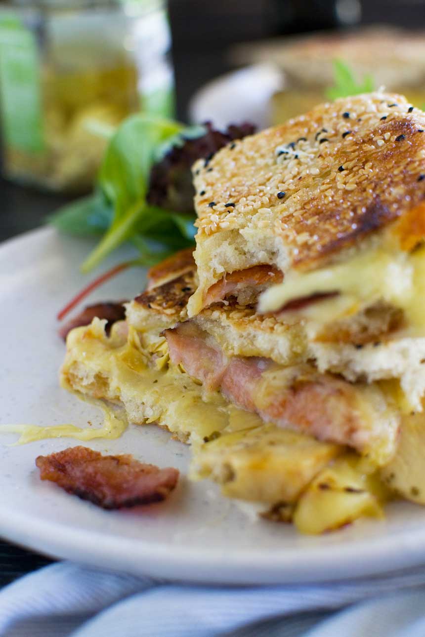 A close up of an artichoke and bacon grilled cheese sandwich on a white plate with green salad in the background.