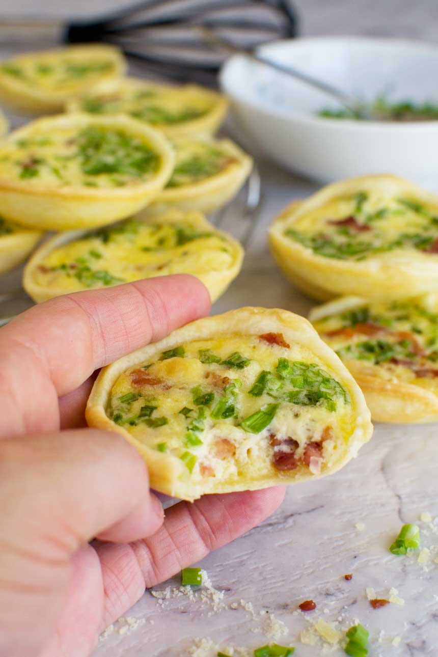 Someone holding up a mini quiche with a bite out of it with others in the background.