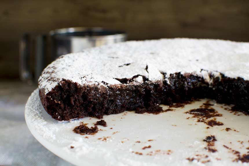 A whole half of a 6-ingredient gooey Swedish chocolate cake  or kladdkaka on a white cake stand and with the gooey middle showing.