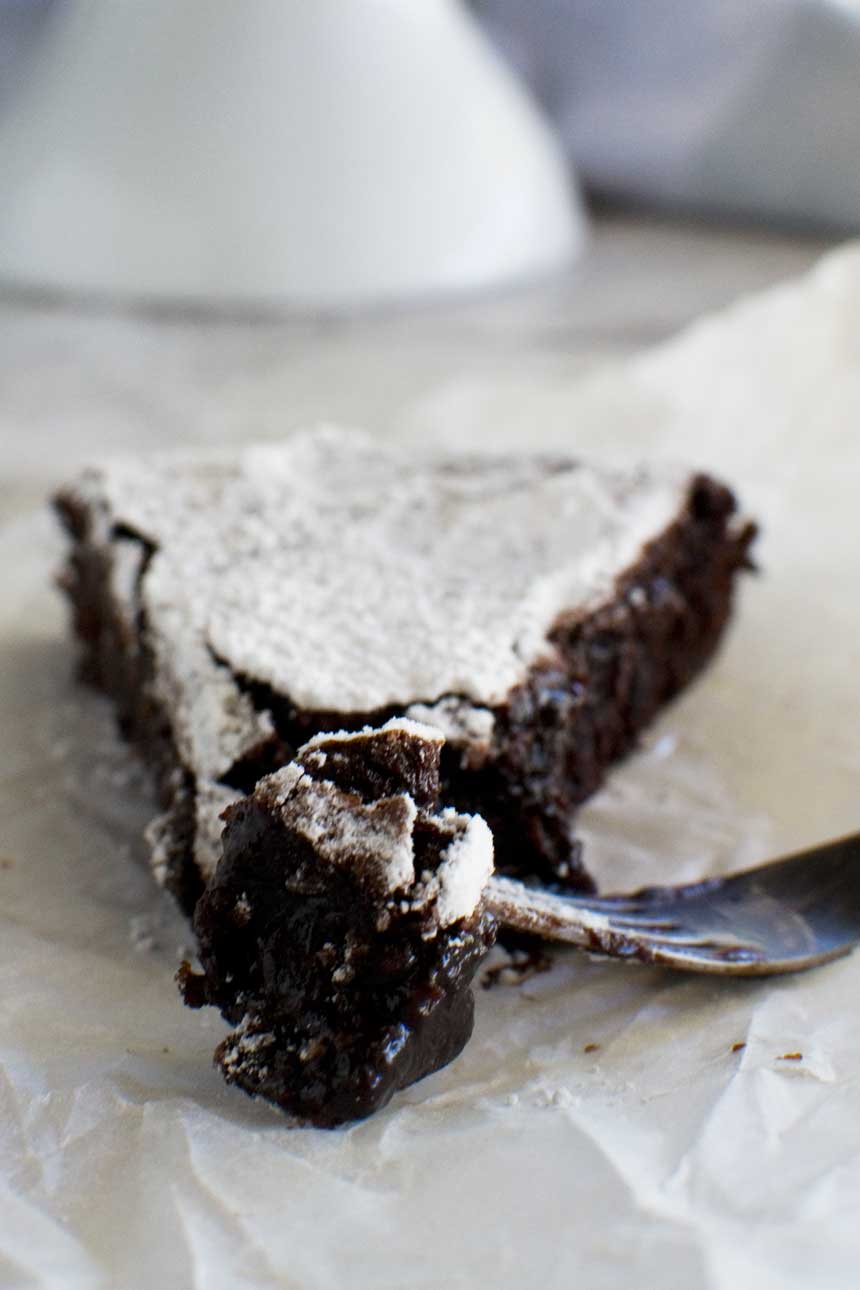 A close-up of a piece of 6-ingredient gooey Swedish chocolate cake or kladdkaka with a fork on baking paper.