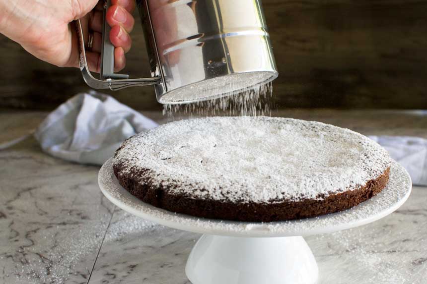 A whole 6-ingredient gooey Swedish chocolate cake on a white cake stand with someone sprinkling icing sugar over it.