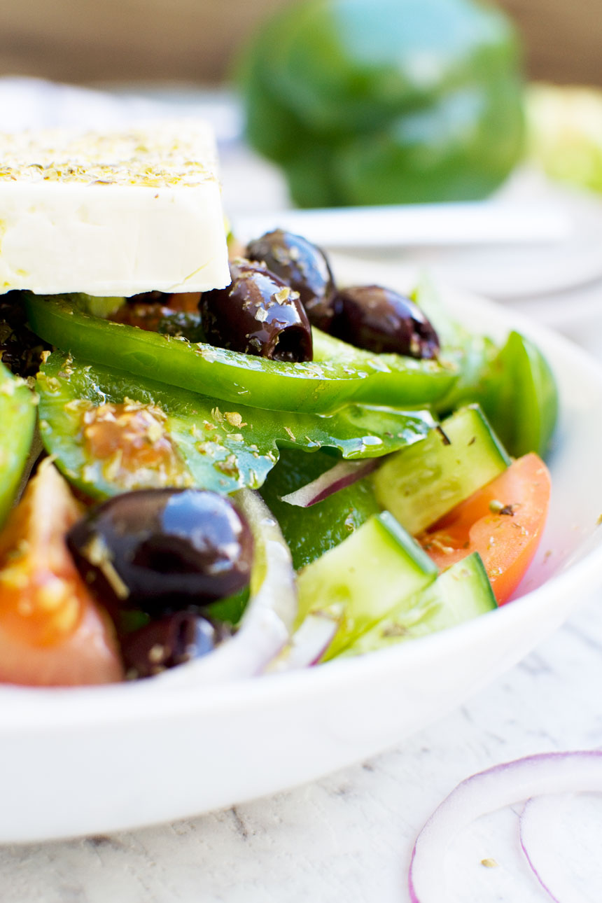 Closeup of a Greek salad in a white bowl with a big block of feta on top and on a marble background.