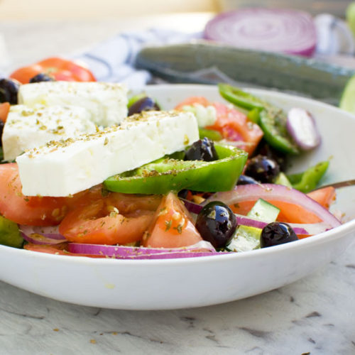 A traditional Greek salad in a white bowl on a marble background and with ingredients behind the bowl.