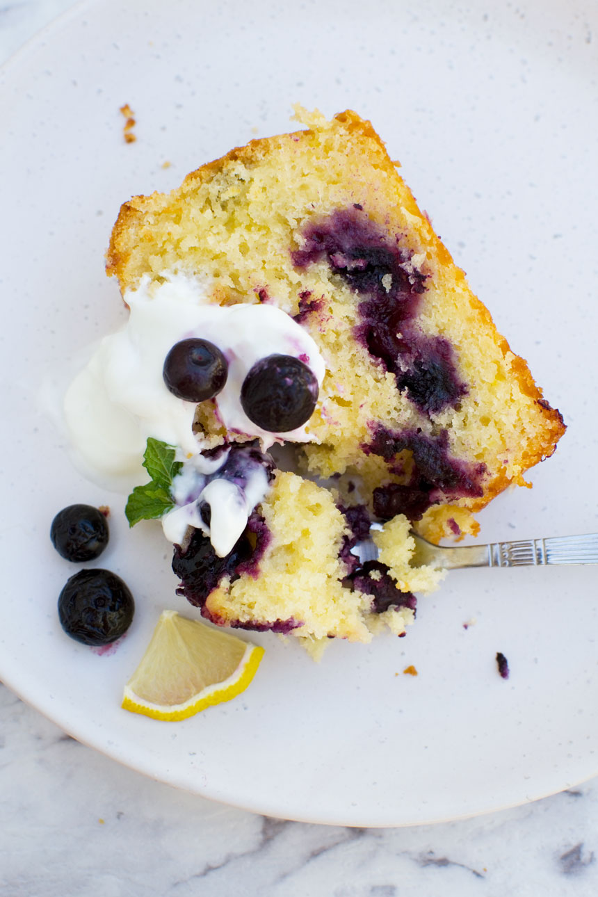 Someone eating a slice of sticky blueberry lemon curd cake with yogurt and fresh blueberries on top on a white plate with a fork from above.