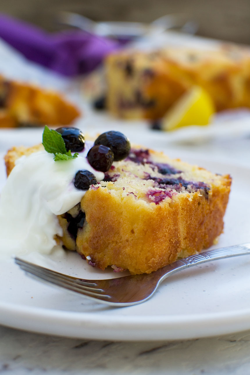 A slice of sticky blueberry lemon curd cake with yogurt and fresh blueberries on on a white plate with a fork and with the rest of the cake in the background.